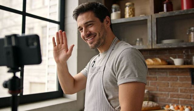 Friendly baker waving during live baking stream in sunlit loft kitchen
