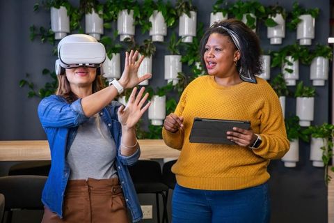Diverse Women Testing Virtual Reality Headset in Office with Mentor Guiding on Tablet