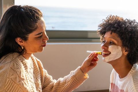 Diverse friends applying facial mask by sunlit coastal window, cozy skincare moment