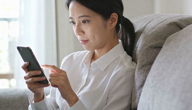 Asian woman scrolling smartphone on sofa holding phone and focusing in natural daylight