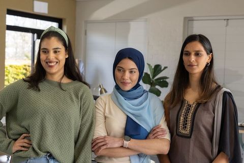 Diverse women standing together at home wearing hijab and kurta, teamwork and friendship