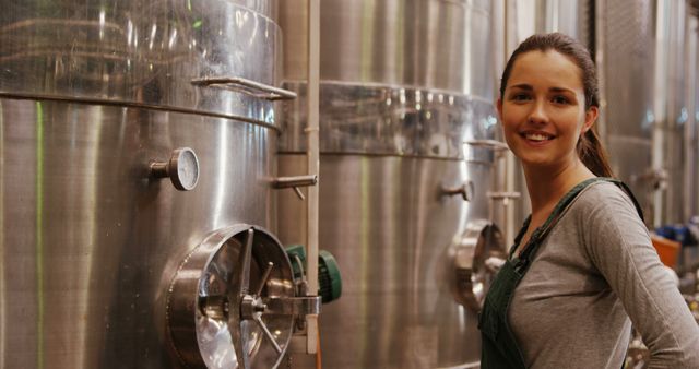 Woman brewer in working environment at craft brewery, standing next to stainless steel fermentation tanks. Useful for articles or advertisements about the brewing industry, female professionals, and craft beer production.