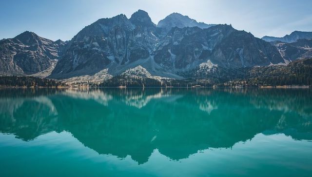 Turquoise alpine lake reflecting jagged mountain peaks and forested shoreline at dawn