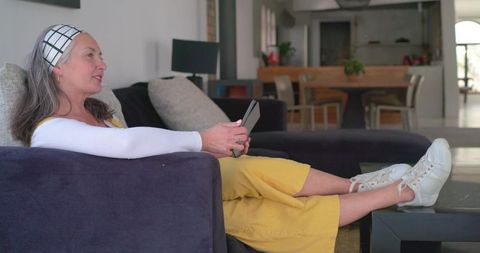 Reclining senior woman using tablet on dark sofa, relaxed lifestyle in open-plan living