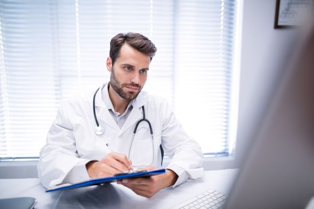 Male doctor writing on clipboard while working on personal computer in clinic