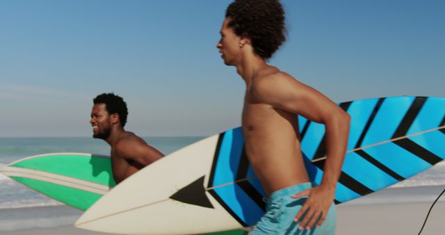 Two young male surfers with curly hair are carrying surfboards while walking on a sandy beach. Both surfers are shirtless and wearing swim trunks. The scene reflects a vibrant and fun beach day under a clear blue sky. This image is ideal for use in advertisements for surfing gear, travel brochures for beach destinations, fitness and lifestyle promotions, or any content related to summer and outdoor activities.