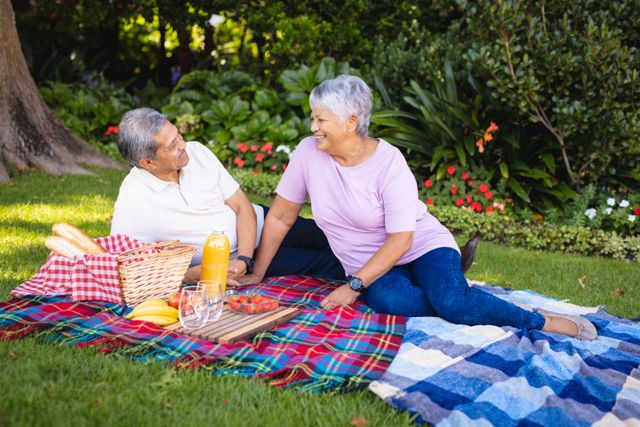 Biracial happy senior couple looking at each other while relaxing on blankets against plants in park. nature, unaltered, love, togetherness, fruit, juice, picnic, lifestyle and retirement concept.
