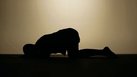 Solitary kneeling silhouette lowering forehead to mat under warm backlit halo yoga pose