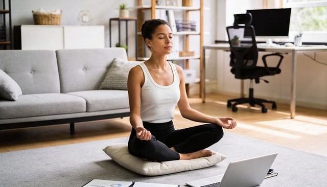 Woman meditating in home office on cushion following online yoga and mindfulness routine