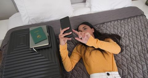 Woman lying on bed scrolling phone beside suitcase, passport, notebook for travel planning