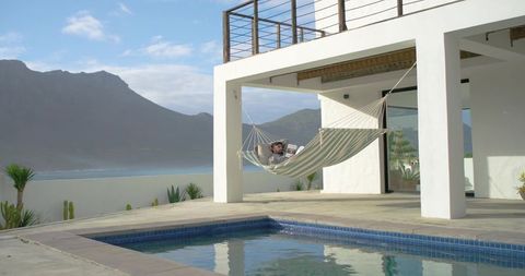 Indian man relaxing in hammock by villa pool with coastal ocean view and mountain backdrop
