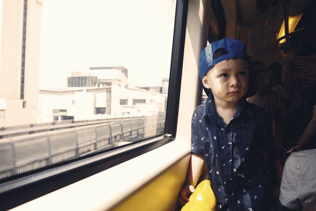 Young boy with a blue hat gazing out the window of a moving commuter train, urban setting visible outside. Perfect for illustrating topics related to city travel, public transportation, childhood, daily commute, urban living, or reflective moments during journey.