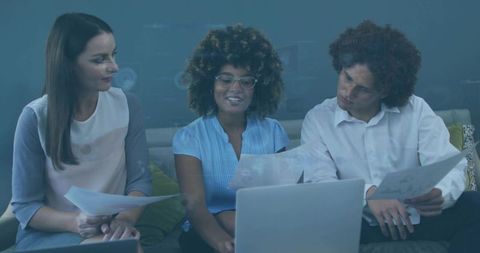 Three coworkers collaborating over laptop while reviewing printed documents on sofa