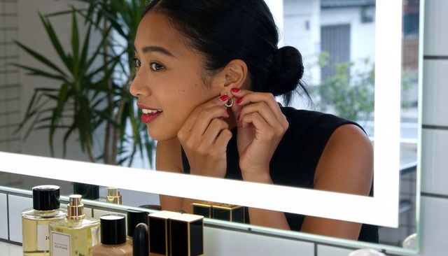 Woman fastening hoop earring at led vanity mirror with perfume bottles and cosmetics