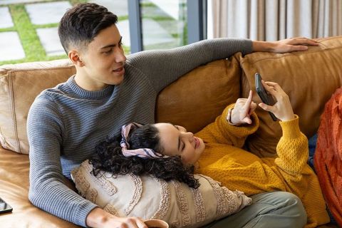 Young couple relaxing on tan leather sofa using smartphone and drinking coffee at home