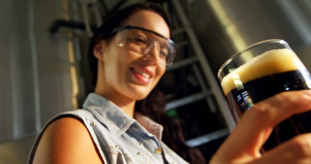 A young Caucasian woman wearing safety glasses is smiling as she holds a glass of dark beer, with copy space. Her attire and the stainless steel tanks in the background suggest she may be a brewmaster or involved in the craft beer industry.