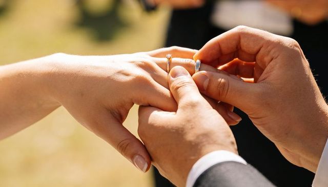 Groom sliding wedding band onto bride's finger during sunlit outdoor ceremony