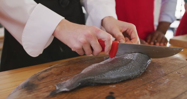 Hands of diverse female cook slicing fish on cutting board and advising senior group in kitchen. Lifestyle, food, cooking and senior lifestyle, unaltered.