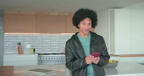 Young man holding smartphone at kitchen island wearing leather jacket and casual tee