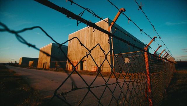 Rusty barbed wire fence guarding wooden warehouses at sunset