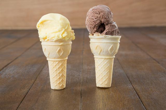 Two ice cream cones, one with chocolate ice cream and the other with vanilla ice cream, are placed on a wooden table. This image can be used for food blogs, summer-themed advertisements, dessert menus, or social media posts promoting sweet treats and refreshments.