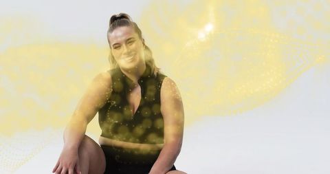 Smiling fit woman sitting in black workout set with ethereal yellow bokeh lights