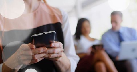 Mature woman holding smartphone in sunlit lounge with colleagues using tablet and laptop