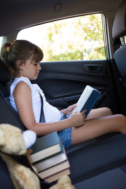 Teenage girl attentively reading a book while sitting in the back seat of a car. Ideal for use in educational materials, travel blogs, family lifestyle articles, and advertisements promoting reading and learning during travel.