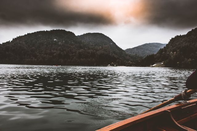 Picture features a wooden rowboat on a tranquil lake, surrounded by dramatic mountain ranges under an overcast sky. Ideal for travel brochures, outdoor adventure promotions, nature-themed posters, or relaxation visuals. An excellent choice for conveying peace, tranquility, and exploration.