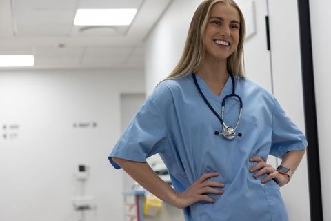 Confident Female Nurse in Blue Scrubs Smiling in Clinic Corridor Wearing Stethoscope