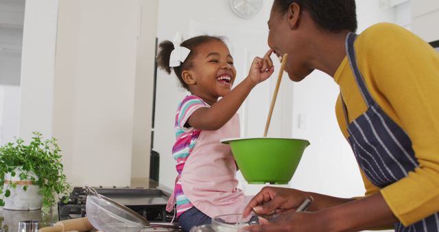 This image captures a joyful moment between a mother and her young daughter baking together in a kitchen. The mother is playfully letting her daughter taste the mixture from a green bowl while both of them smile and enjoy the activity. Can be used for promoting family bonding activities, parenting articles, home baking kits, and cooking classes. Highlights the importance of spending quality time and creating memories at home.
