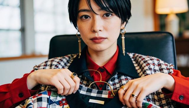 Confident stylist presenting patterned coat on hanger wearing red blouse and bold jewelry