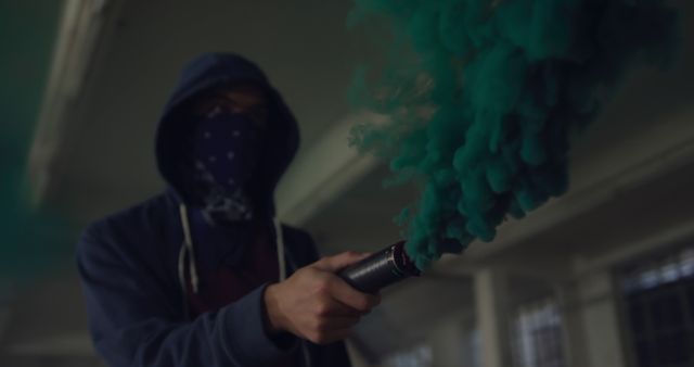 Mysterious hooded person holds a smoke canister emitting green smoke in an indoor setting. The individual wears a mask, adding an element of anonymity. Suitable for use in themes related to mystery, atmosphere, urban, rebellion, or hidden identity.