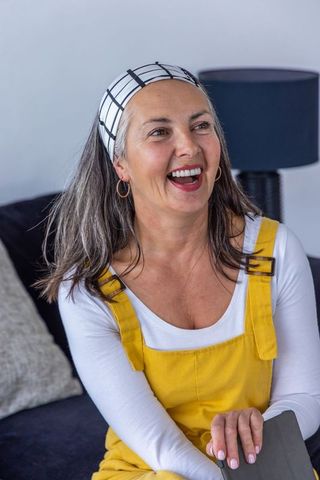 Mature woman laughing and glancing left on sofa wearing yellow overalls and headband