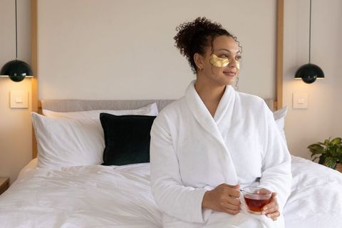 African american woman relaxing in robe with gold under-eye patches sipping tea on bed