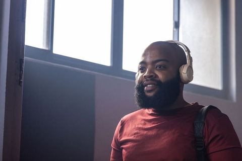 Bearded man listening to music with headphones in sunlit lobby wearing backpack, reflective