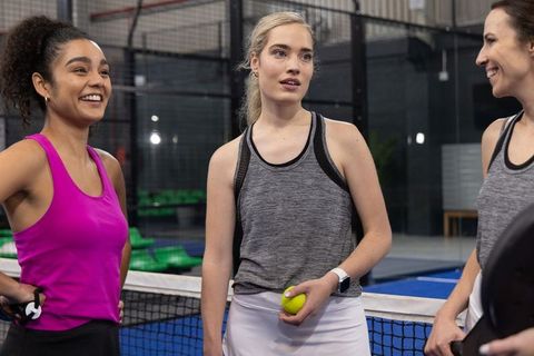 Diverse women chatting on indoor padel court wearing sporty tank tops and holding ball