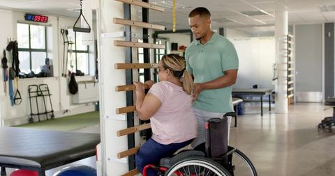 Therapist guiding wheelchair user through stall-bar upper-body exercise in rehab clinic