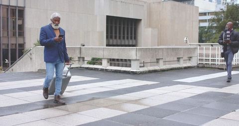 Mature African American Man Walking with Rolling Suitcase Checking Phone in Urban Plaza