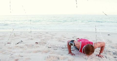 Young man performing push-ups on sunlit sandy beach wearing smartphone armband workout