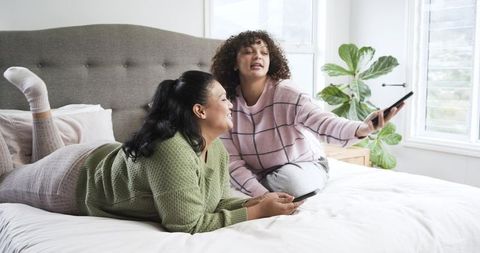 Mother and teen daughter lounging on bed in sunlit bedroom holding smartphones and remote, cozy morn
