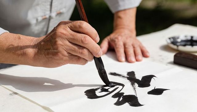 Hands of calligrapher creating bold black ink strokes with traditional brush in sunlight
