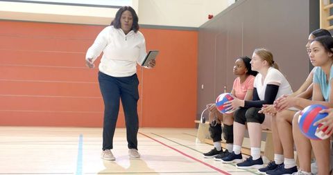 Mature african american coach coaching female volleyball players on bench with tablet