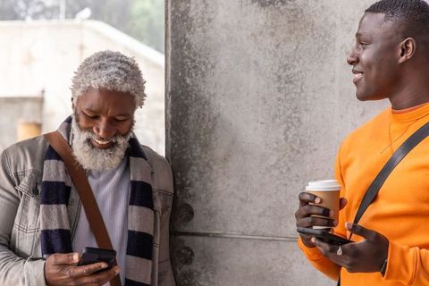 Men sharing smartphone laugh while enjoying coffee beside concrete pillar urban moment