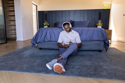 African man relaxing on bedroom rug using tablet and stylus with headphones, modern calm