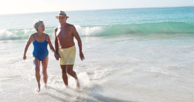 Elderly couple holding hands and walking along the shoreline with gentle waves. They are wearing swimwear suitable for a beach day. This image is ideal for promoting retirement communities, travel agencies specializing in senior holidays, and wellness activities for seniors. It evokes feelings of companionship, relaxation and an active retired life.