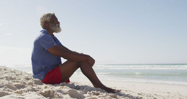 Senior man sitting on the beach during a sunny day, clad in red shorts and a blue shirt, gazing at the ocean with a serene expression. Ideal for themes of relaxation, retirement, leisure, senior lifestyle, seaside vacations, and peaceful moments. Can be used in travel brochures, wellness magazines, advertisements for travel agencies, or articles on mental well-being and relaxation.