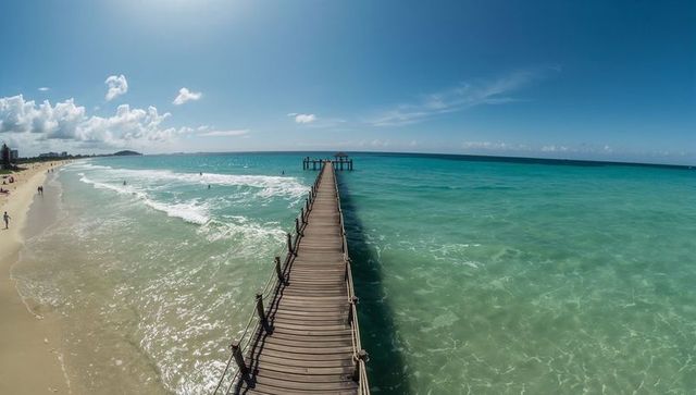 Wooden pier stretching toward horizon over turquoise ocean with sandy beach and swimmers