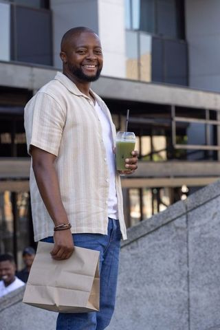 Smiling african man holding matcha smoothie and paper bag on urban stone steps
