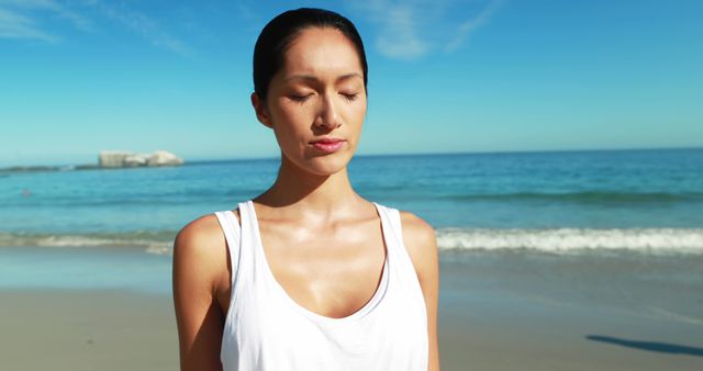 Young woman meditating on sunny beach with eyes closed. Perfect image for wellness campaigns, meditation apps, travel promotions, or health and mindfulness blogs. The calm ocean and clear blue sky enhance the peaceful ambiance.